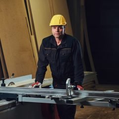 Carpenter male dressed in a safety yellow cap works with chain-saw in a garage.